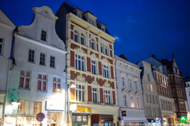 Historic Lübeck townhouses at blue hour, including an ornate red-and-cream gabled façade with tall grouped windows and illuminated shops.