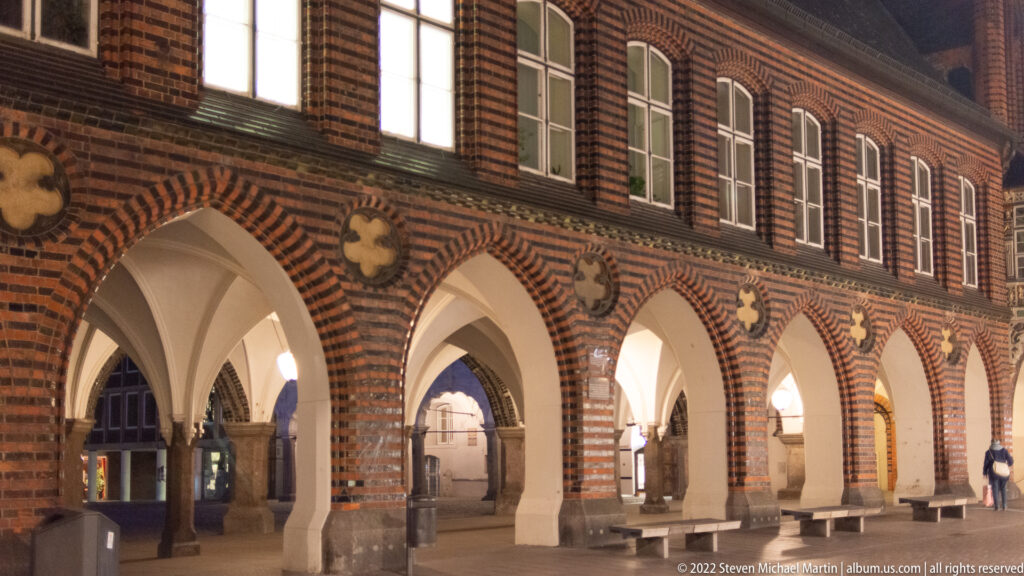 Illuminated pointed brick arcades beneath Lübeck Town Hall, with pale vaults, patterned masonry, and circular decorative insets.