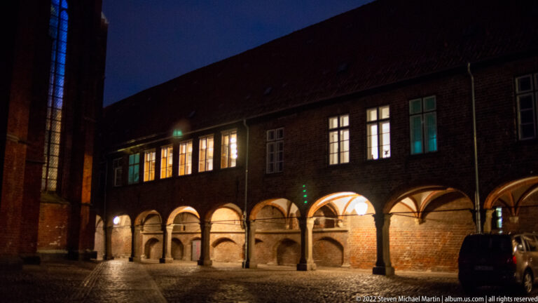 Night view of a long historic brick arcade in Lübeck, with illuminated round arches, cobblestones, and simple upper-story windows.