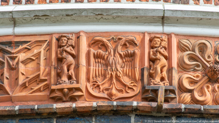Terracotta relief on the Holstentor with a central eagle, small standing figures, and scrolling ornament in dark brickwork.