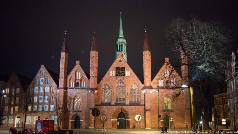 Illuminated main façade of Lübeck’s Heiligen-Geist-Hospital at Koberg, with pointed gable, tall windows, four slender turrets, and green spire.