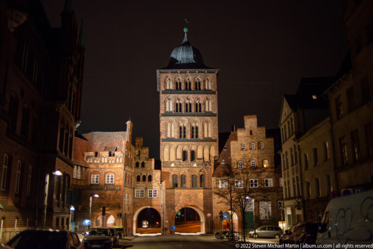 Illuminated Burgtor in Lübeck at night, with tall brick tower, stacked arcades, arched gateway, and adjoining stepped-gable buildings.