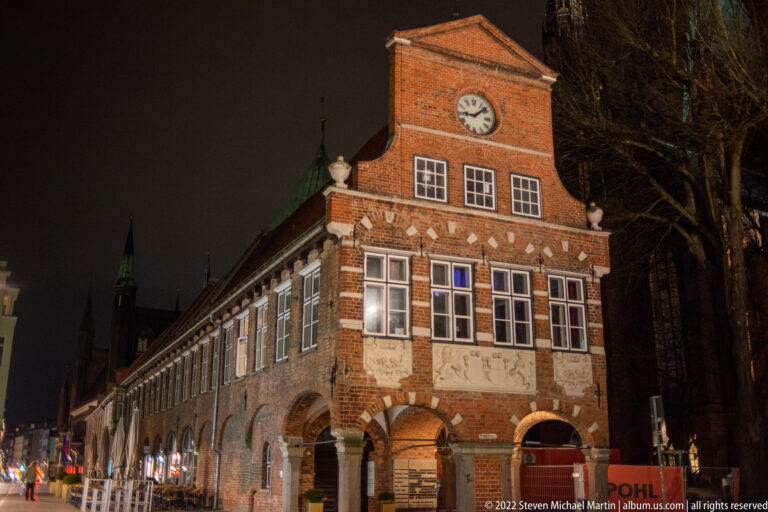 Night view of the Heiligen-Geist-Hospital’s clock-gabled brick wing with repeated ground-floor arcade and white-framed windows.