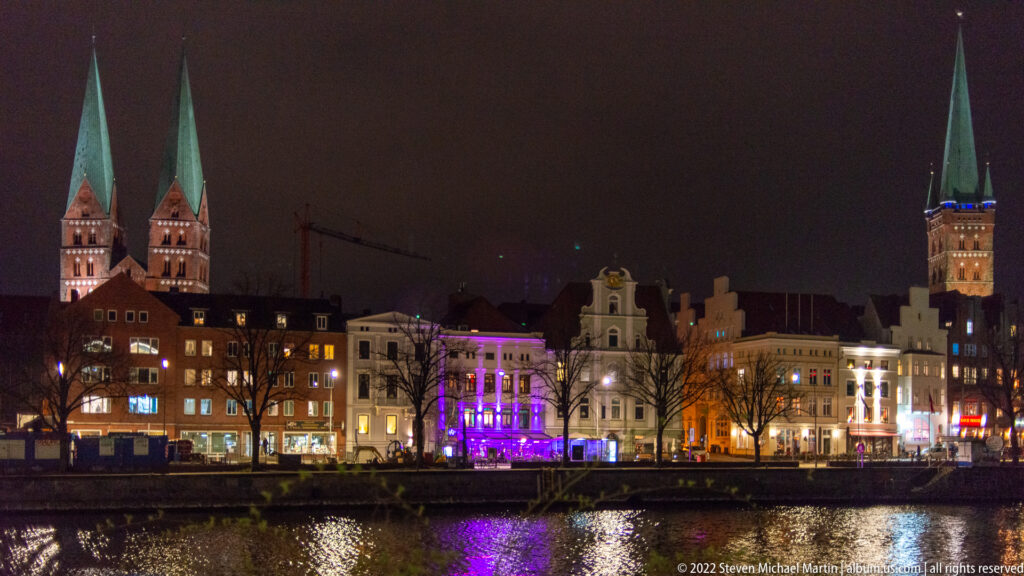 Night skyline of Lübeck across the Trave, with illuminated waterfront façades, reflected lights, and multiple green-spired church towers.