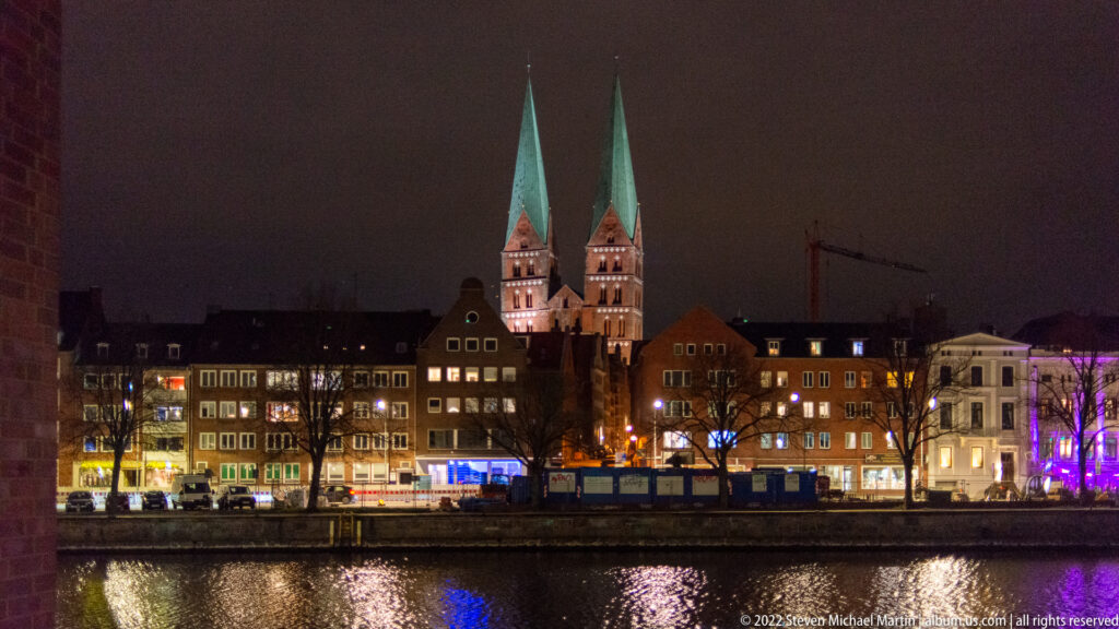 Twin illuminated brick church towers rising above Lübeck’s historic waterfront at night, with green spires and reflections on the Trave.