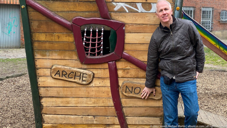Visitor beside a colorful Noah’s Ark-themed wooden play structure with timber walls, rope-net window, and painted lettering.