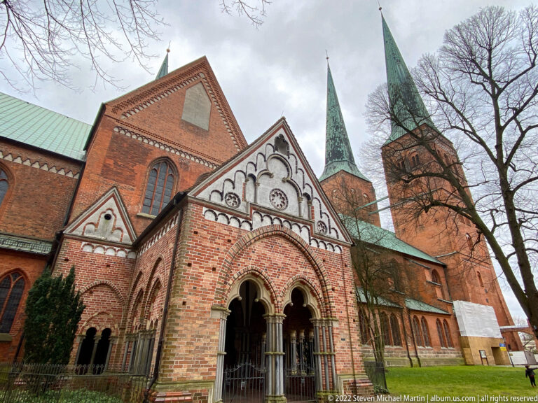 Close view of Lübeck Cathedral’s ornate Paradise porch with rounded arches, pale columns, patterned brickwork, and twin green spires behind.