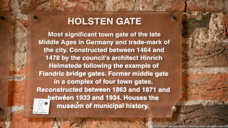 Brown historical plaque labeled “HOLSTEN GATE” mounted on old brickwork, with English text about the gate’s history.