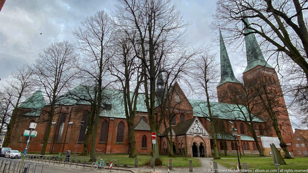Wide exterior view of Lübeck Cathedral with long red-brick nave, green copper roofs, projecting chapels, leafless trees, and twin western towers.