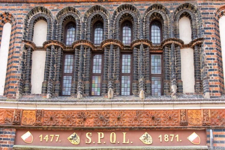 Arched Holstentor windows above terracotta ornament and a band marked “1477 S.P.Q.L. 1871.”