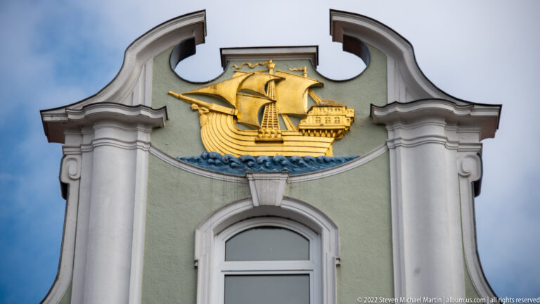 Gilded sailing ship above blue waves on a pale-green curved gable with white architectural trim.