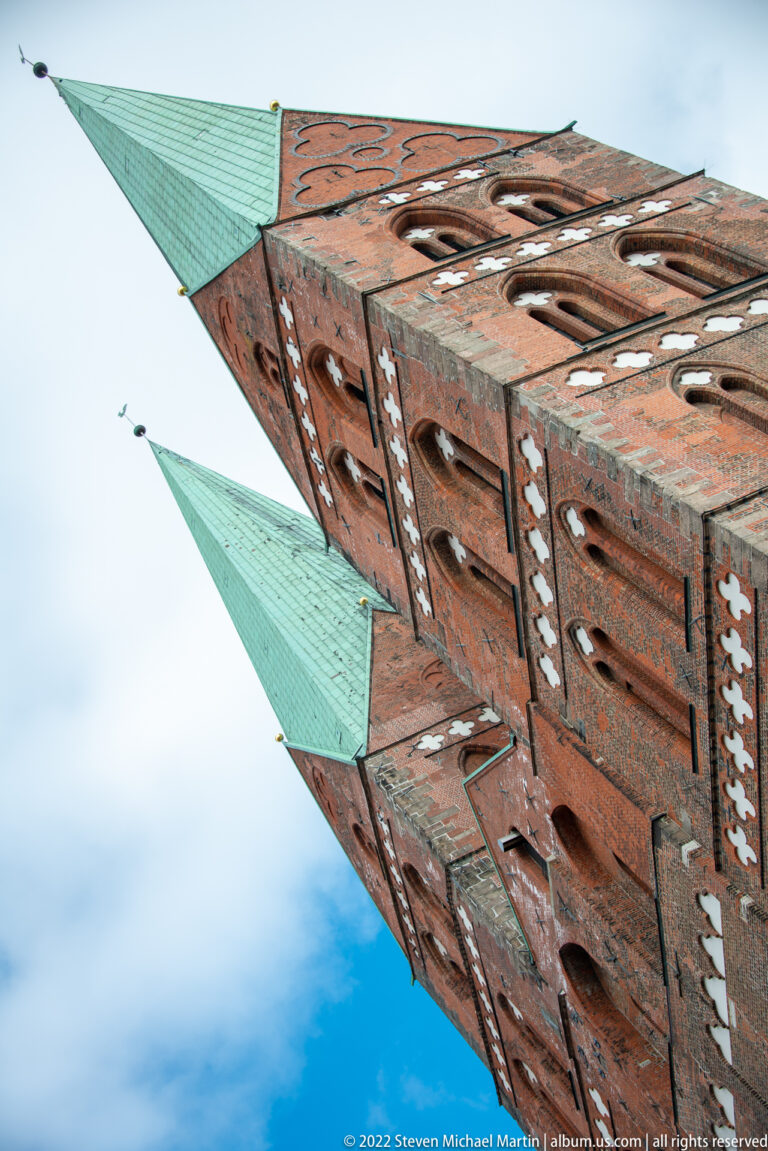 Upward view of two tall red-brick church towers with green copper spires against blue sky.