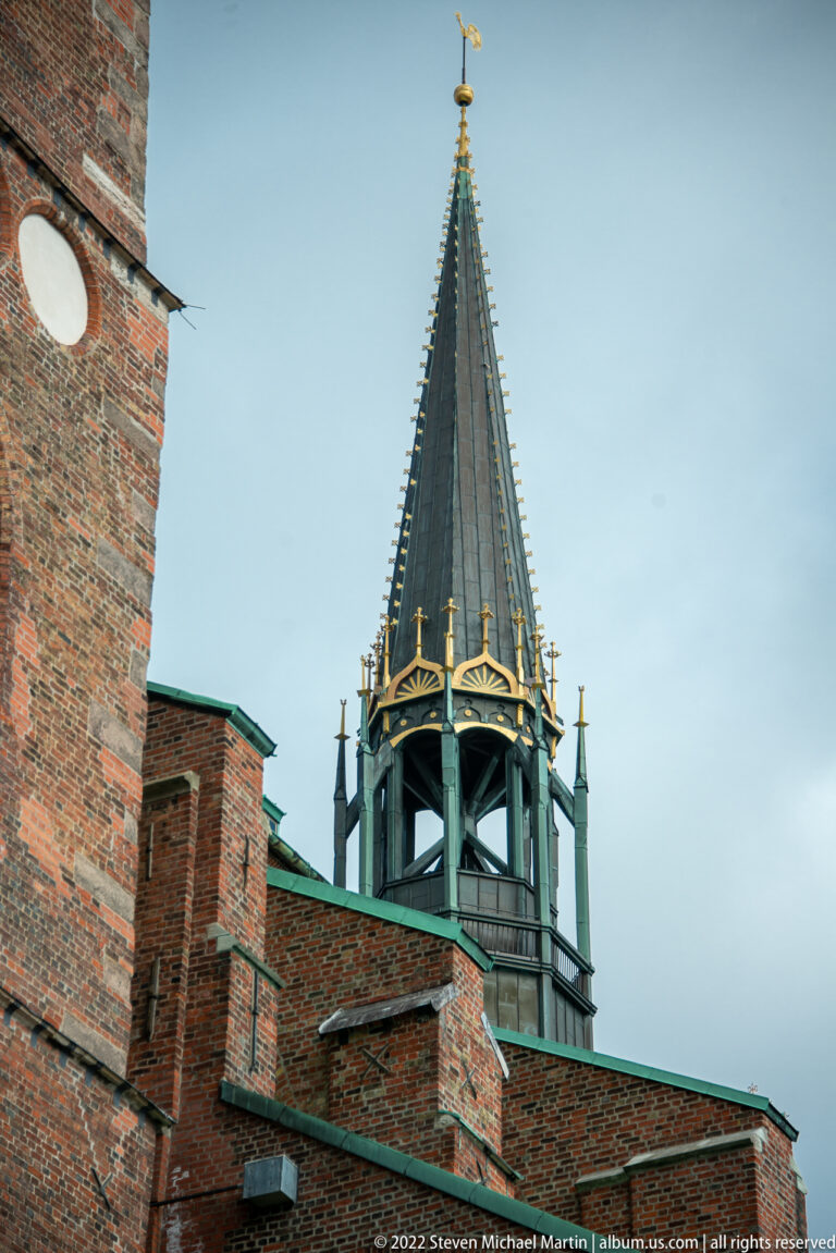 Octagonal open lantern tower with a dark pointed roof, gilded finial, and small corner pinnacles above brick walls.