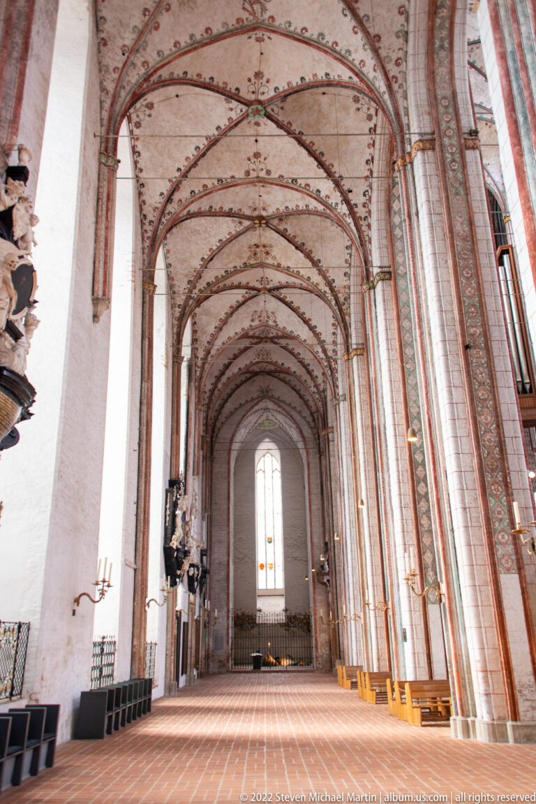 Long Gothic church aisle with red-brick piers, pointed arches, painted ribbed vaults, white walls, and a tall end window.