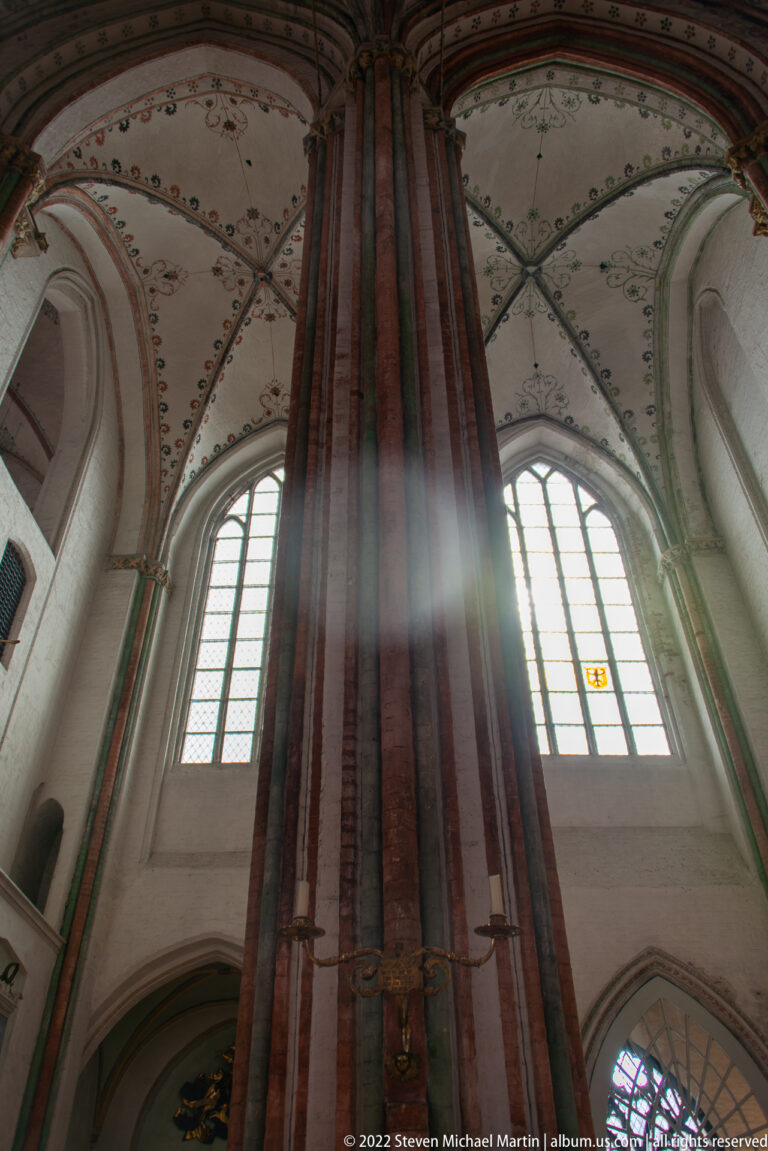 Upward view of a tall brick church pier, arched windows, painted rib vaults, and shafts of light.