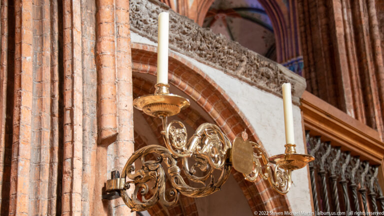 Ornate brass two-candle wall sconce mounted on a red-brick church pier beside carved stonework.