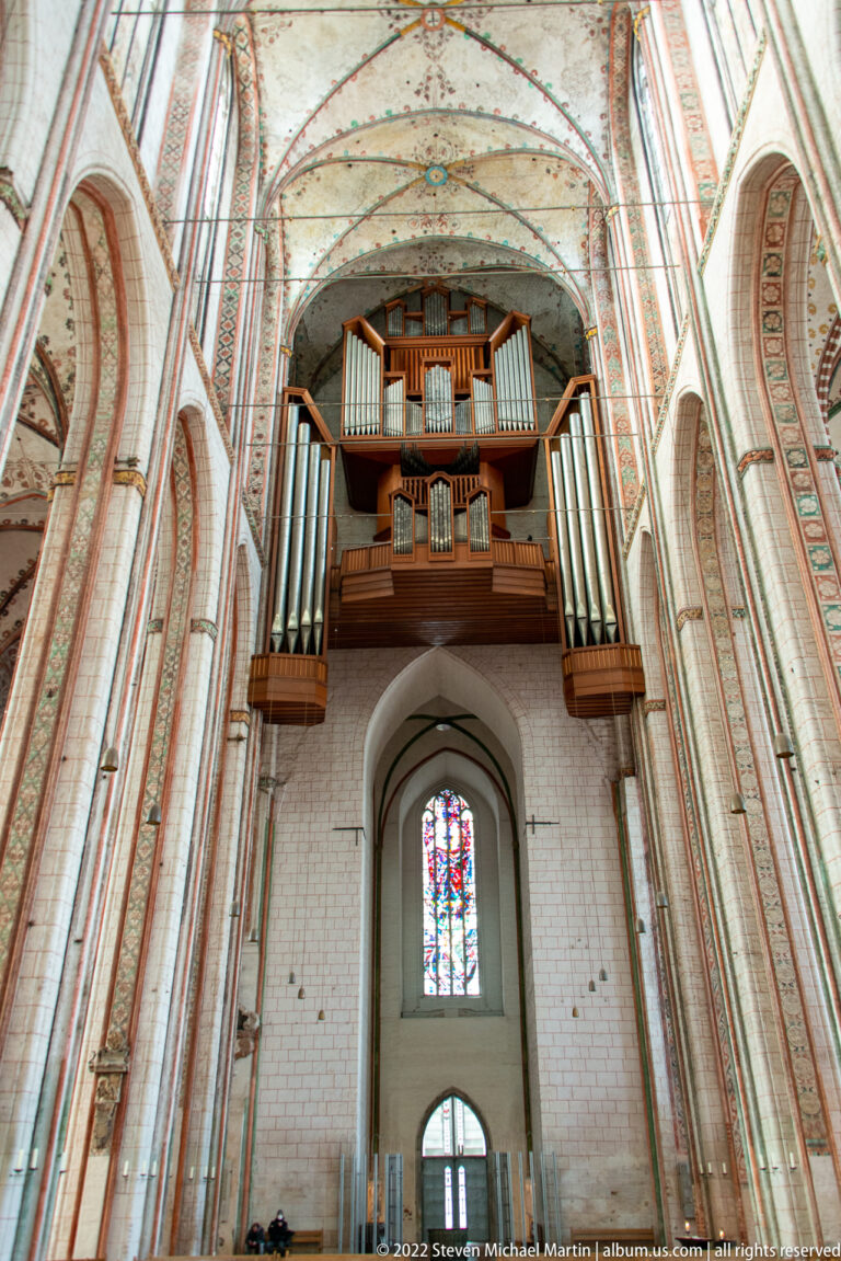 Large multi-section pipe organ above a pointed opening and stained-glass window, framed by brick piers and vaulted ceiling.