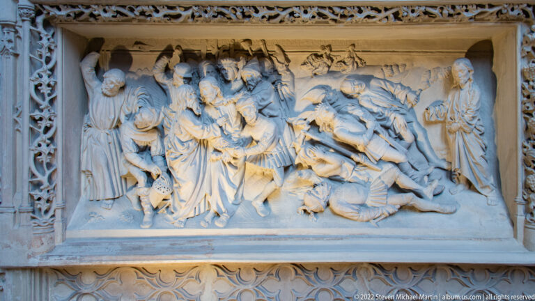 Pale stone relief showing a crowded biblical scene with robed figures around a fallen or kneeling figure, framed by carved ornament.