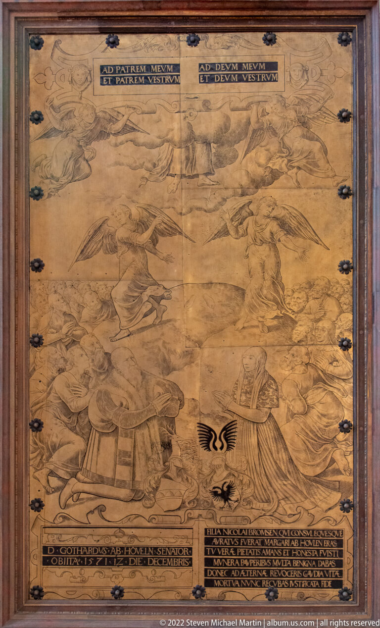 Large framed monochrome church painting with many kneeling and reclining figures, landscape background, and dark inscription bands.