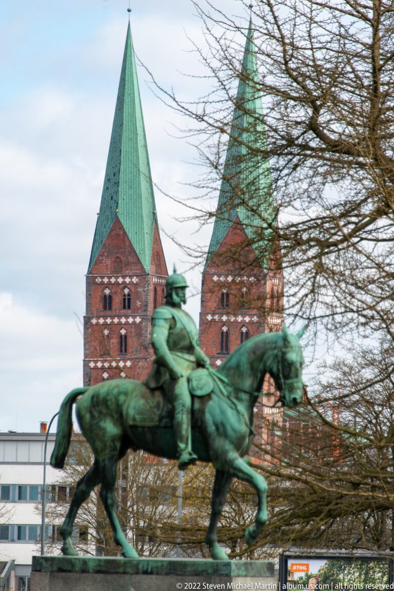 Green bronze equestrian monument before two tall red-brick church towers with copper spires in Lübeck.