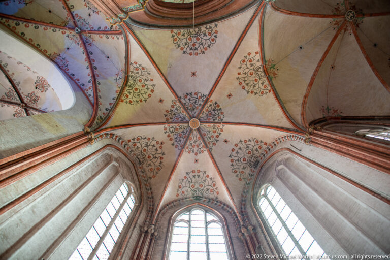 Upward view of a Gothic rib vault with red ribs, pale panels, floral painted ornament, and tall windows below.