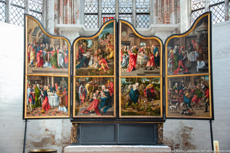 Opened painted winged altarpiece with multiple narrative scenes, dark frames, and leaded church windows behind.