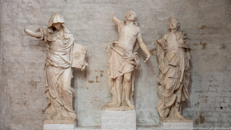 Three pale carved standing figures against a brick church wall, with a partially unclothed central figure.