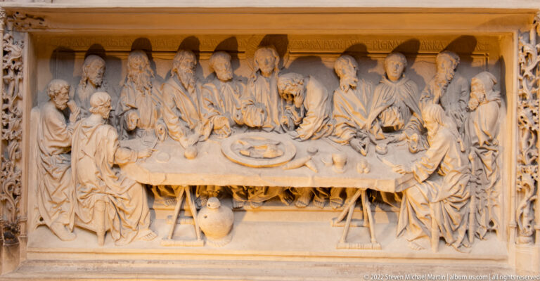 Carved Last Supper relief with Christ and apostles seated around a long table amid vessels and decorative framing.