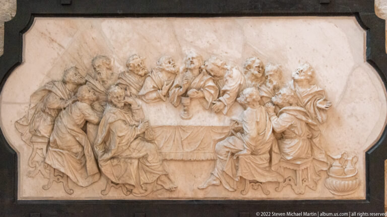 Pale carved relief of many figures gathered around a table, set within a dark-edged shaped frame.