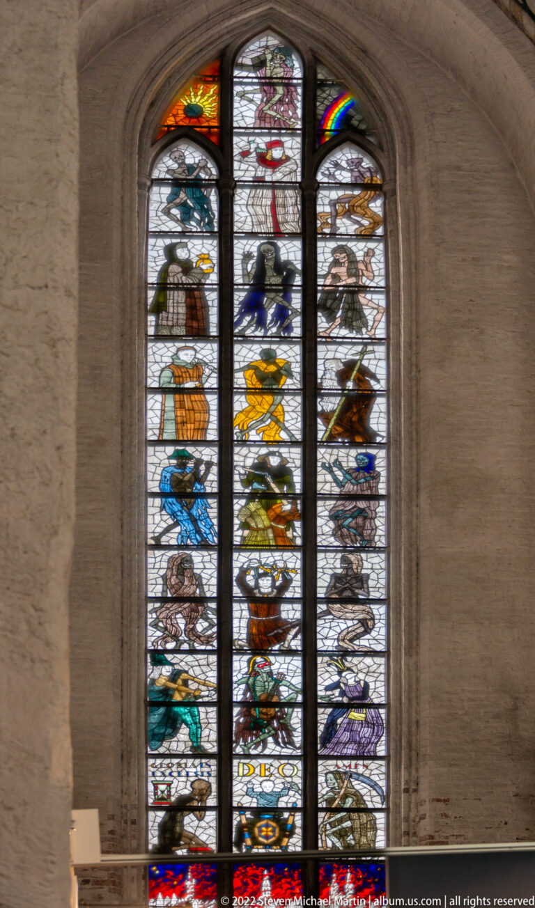 Tall Gothic stained-glass window with heraldic shields, figures, and colorful panels divided by dark tracery.