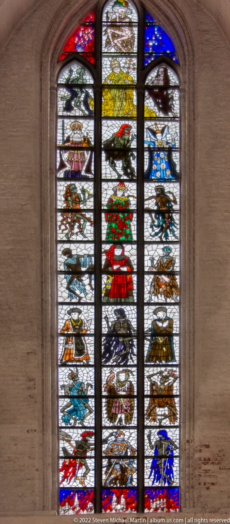 Colorful stained-glass window with shields, animals, costumed figures, and decorative panels in narrow vertical lights.