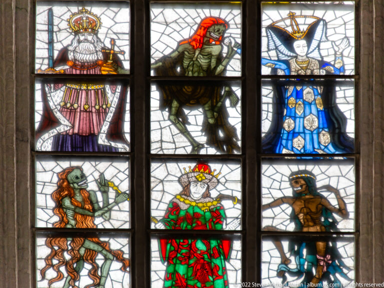 Detail of Alfred Mahlau’s Dance of Death window with richly dressed human figures and skeletal Death figures in vertical stained-glass panels.