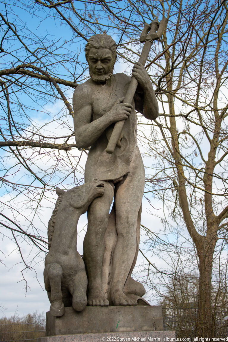 Weathered stone male figure holding a long staff, with a small animal at his leg beneath leafless trees.