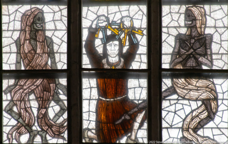 Crowned ruler with raised hands flanked by two shrouded Death figures in Alfred Mahlau’s Lübeck Dance of Death stained glass.