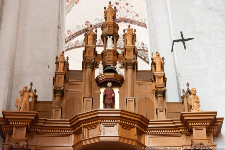Elaborate carved wooden organ case or gallery with columns, statues, and a central elevated figure inside a brick church.