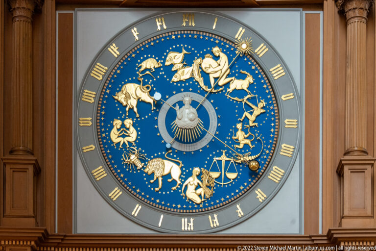 Blue circular astronomical-clock dial with gilded zodiac figures, Roman numerals, and a central celestial motif.