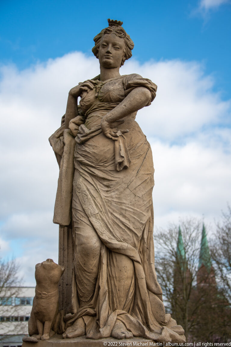 Crowned stone female figure in classical drapery with a small sculptural attribute at her side under blue sky.