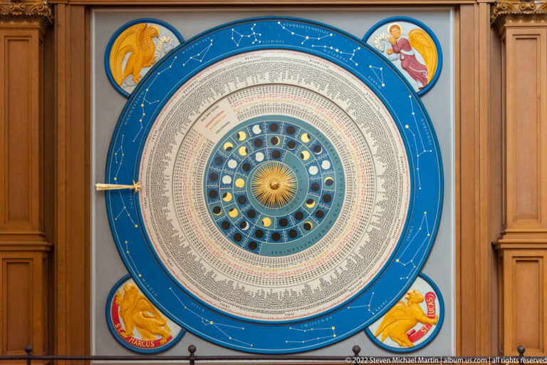 Large blue astronomical calendar dial with concentric text rings, lunar phases, central golden sun, and corner medallions.