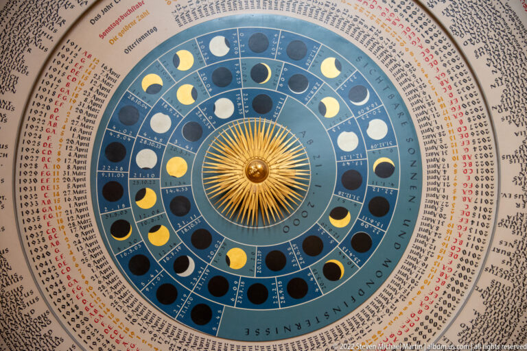 Close view of astronomical clock dial with central golden sun, concentric moon phases, and dense calendar inscriptions.