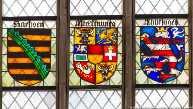 Three colorful coats of arms beneath black-letter inscriptions in a diamond-paned stained-glass window.