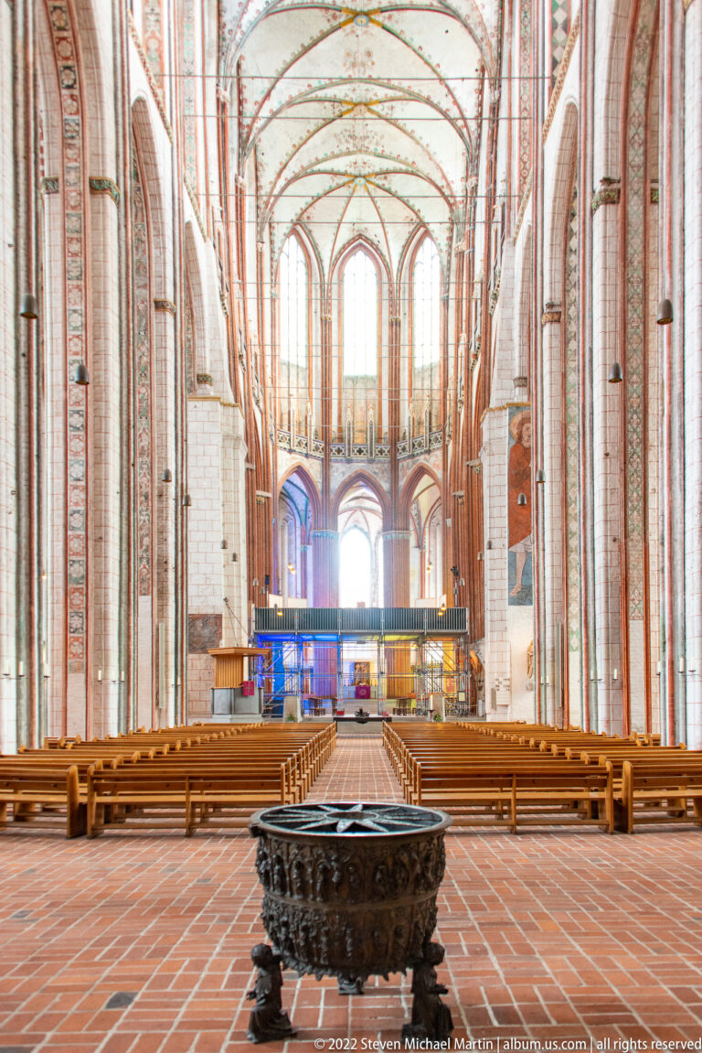 Wide view down the brick Gothic nave with pointed vaults, wooden pews, tall windows, and a dark bronze vessel in front.