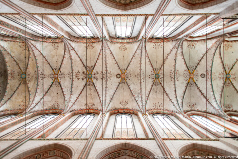 Upward view of pale Gothic vaults with red ribs, small painted ornaments, clerestory windows, and brick supports.