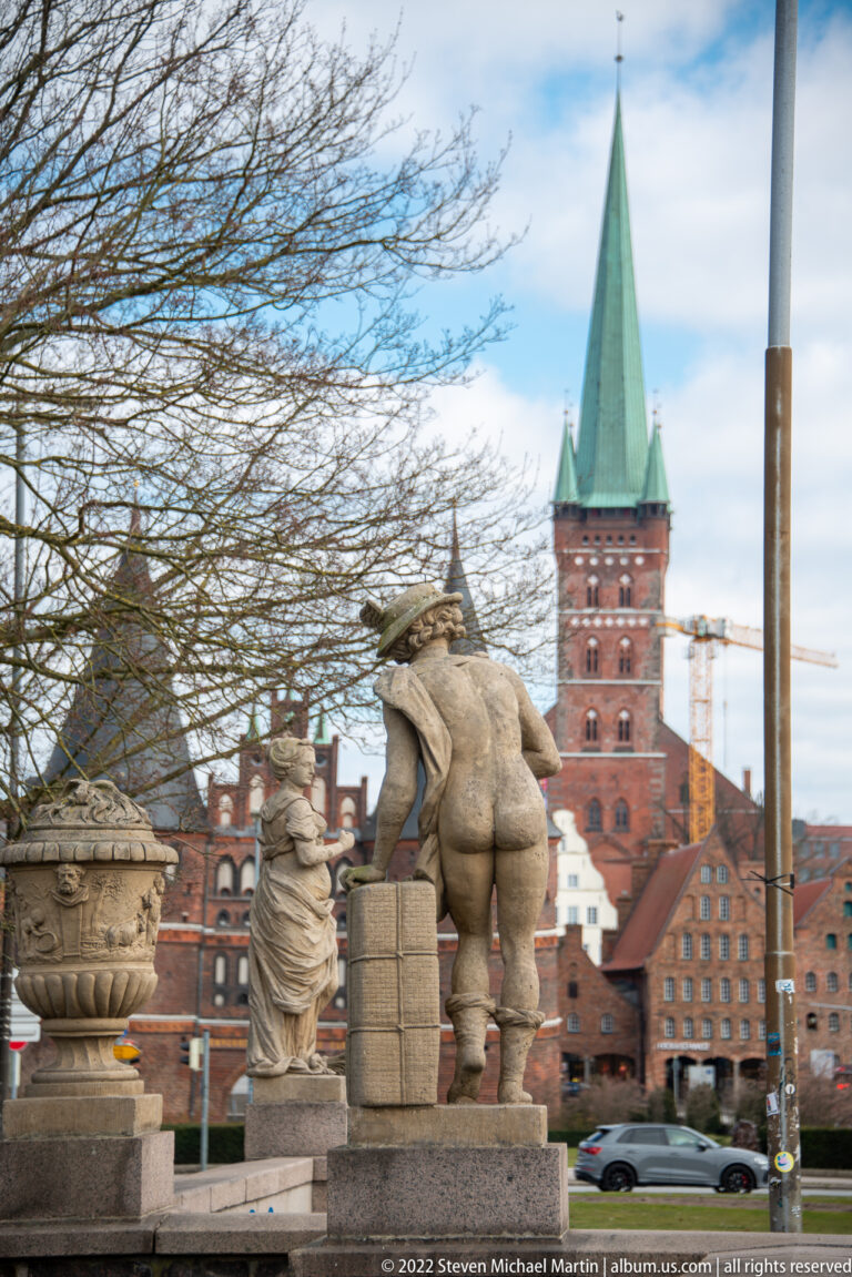 Group of stone statues in an urban space with red-roofed buildings and a tall brick church tower beyond.