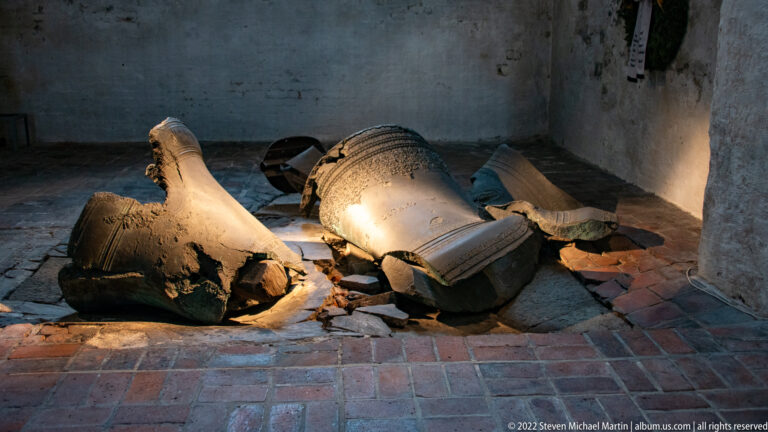 Broken bronze bells and fragments lying on a brick floor in a dark interior corner of St. Mary's Church.
