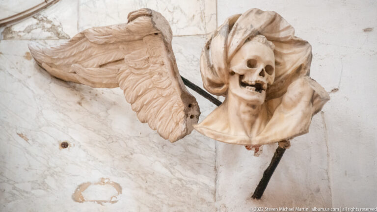 Winged skull sculpture with billowing carved forms mounted high on a pale church wall.