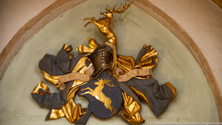 Gilded heraldic display with stag shield, stag crest, dark mantling, and inscribed ribbon beneath a pointed arch.