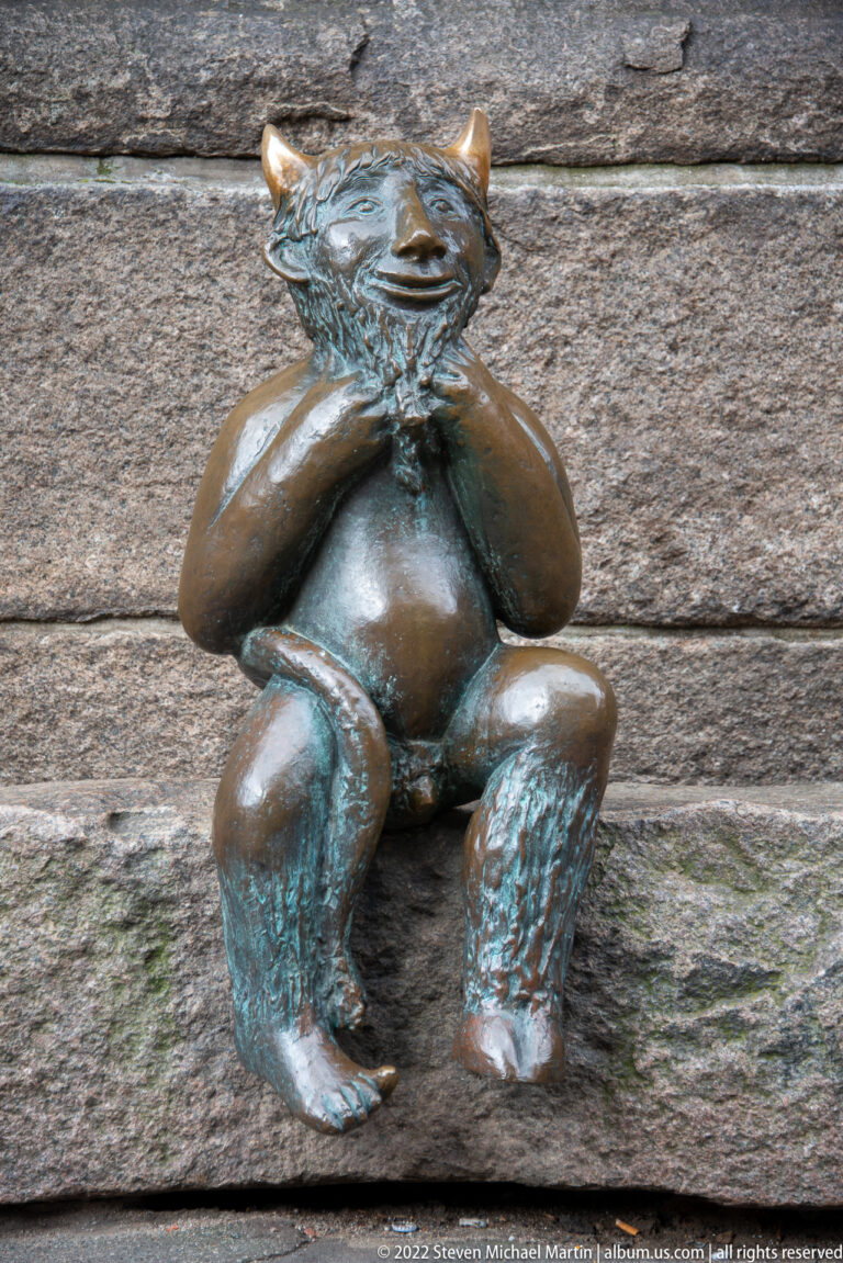 Small seated bronze devil with folded arms, pointed ears, grin, and green patina against a stone wall.