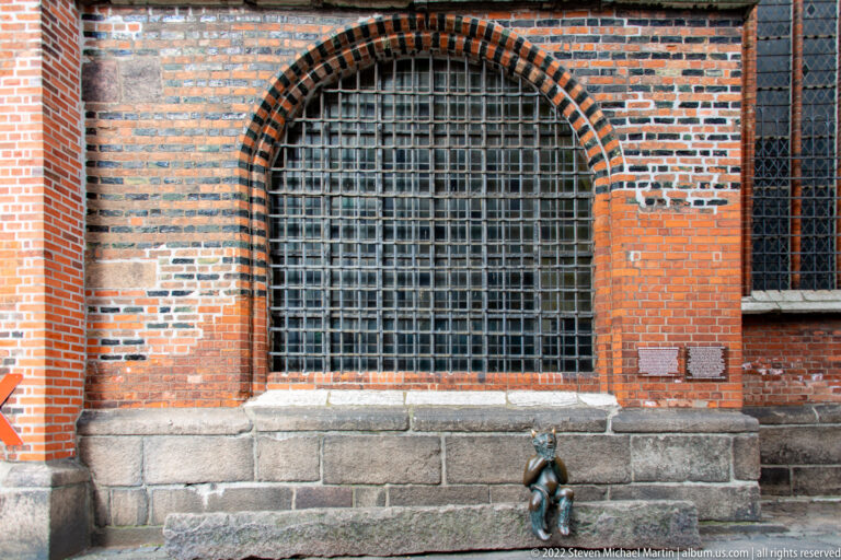 Small bronze devil at the base of a large barred pointed-arch window in striped brick masonry.