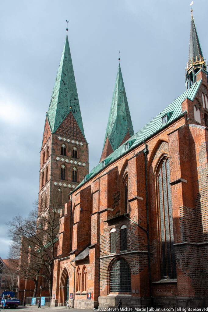 Large Brick Gothic church with twin square towers, green copper roofs, pointed windows, and red-brick nave.