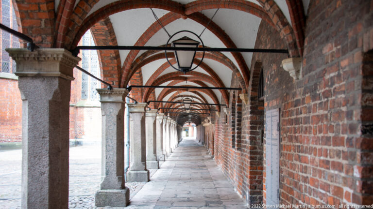 Long vaulted arcade with brick arches, pale stone columns, hanging lanterns, and a paved walkway.